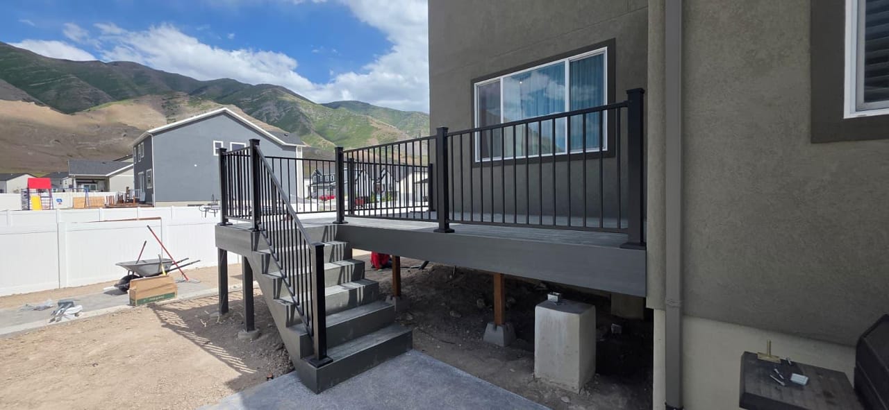 New gray composite deck with black metal railing under construction, stairs to concrete patio, Utah mountains in background — by CasaMás Construction