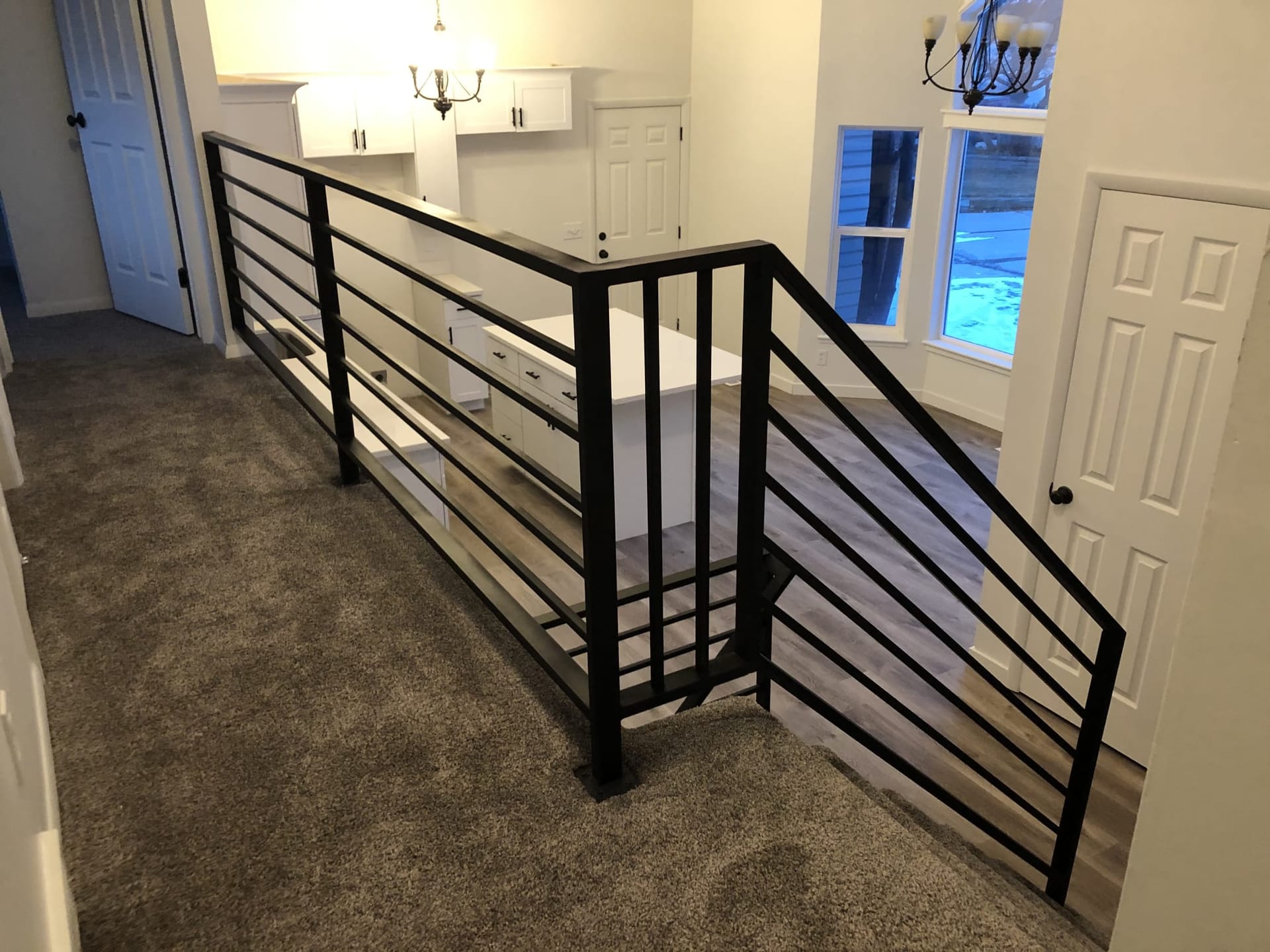 Interior stairwell with custom black horizontal-bar metal railing, hardwood landing, and view to open kitchen with chandelier — Utah interior railing by CasaMás Construction