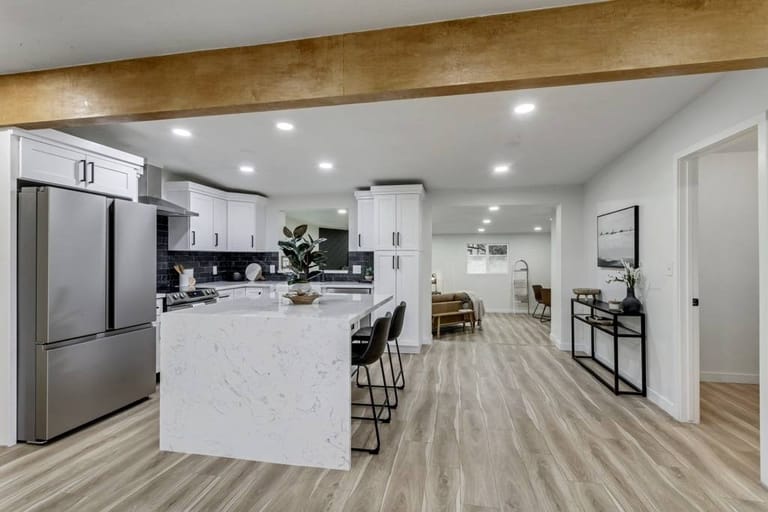 Open-flow kitchen and living area with marble waterfall island, white cabinets, exposed wood beam ceiling, and continuous light-wood plank flooring — Utah open-plan home addition by CasaMás Construction