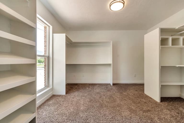 Master walk-in closet with custom built-in shelving on both walls, central hanging unit, carpet, and natural light from window — Utah closet build-out by CasaMás Construction